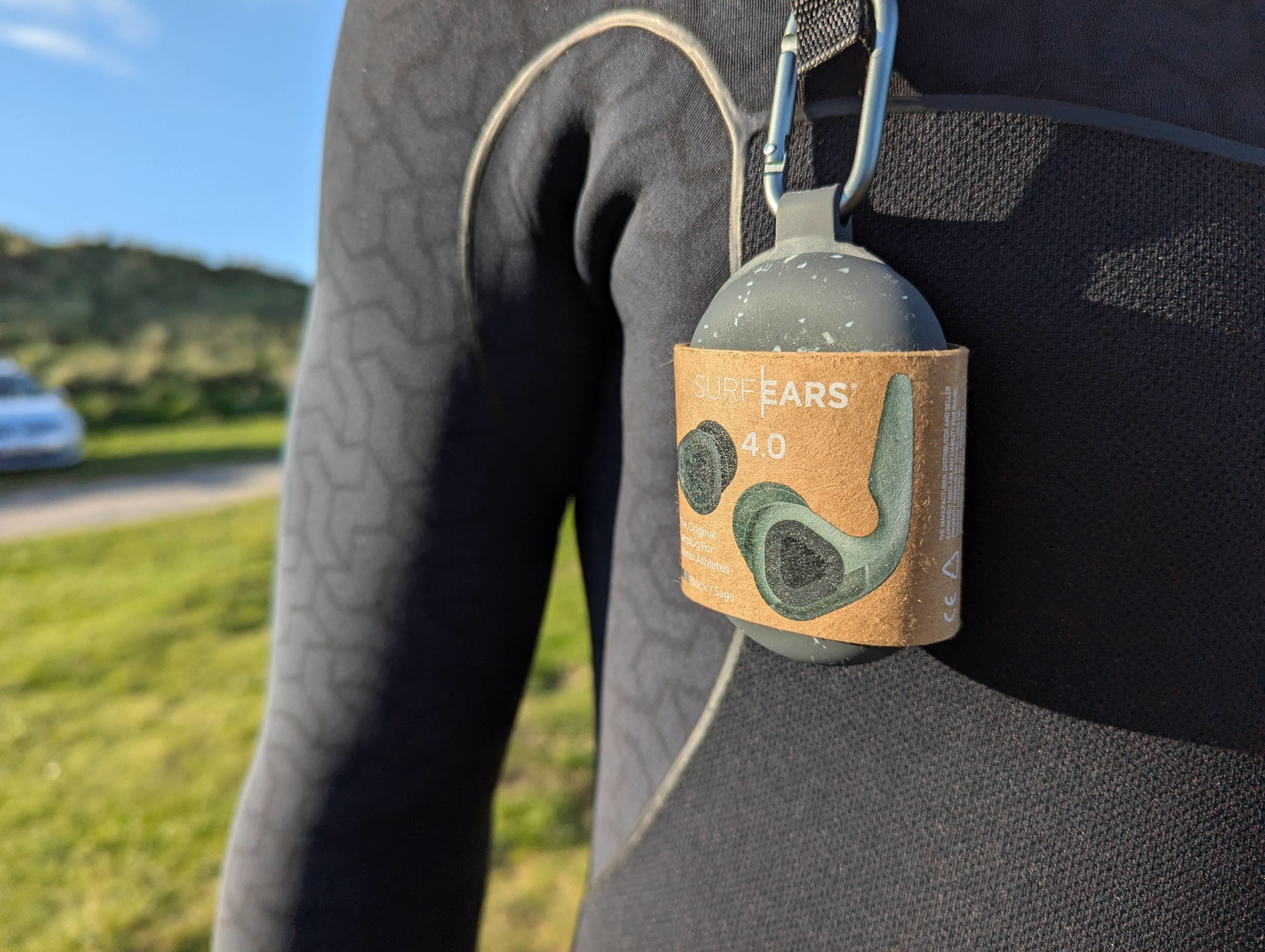 SurfEars earplug case clipped by carabiner to the sleeve of a black wetsuit outdoors