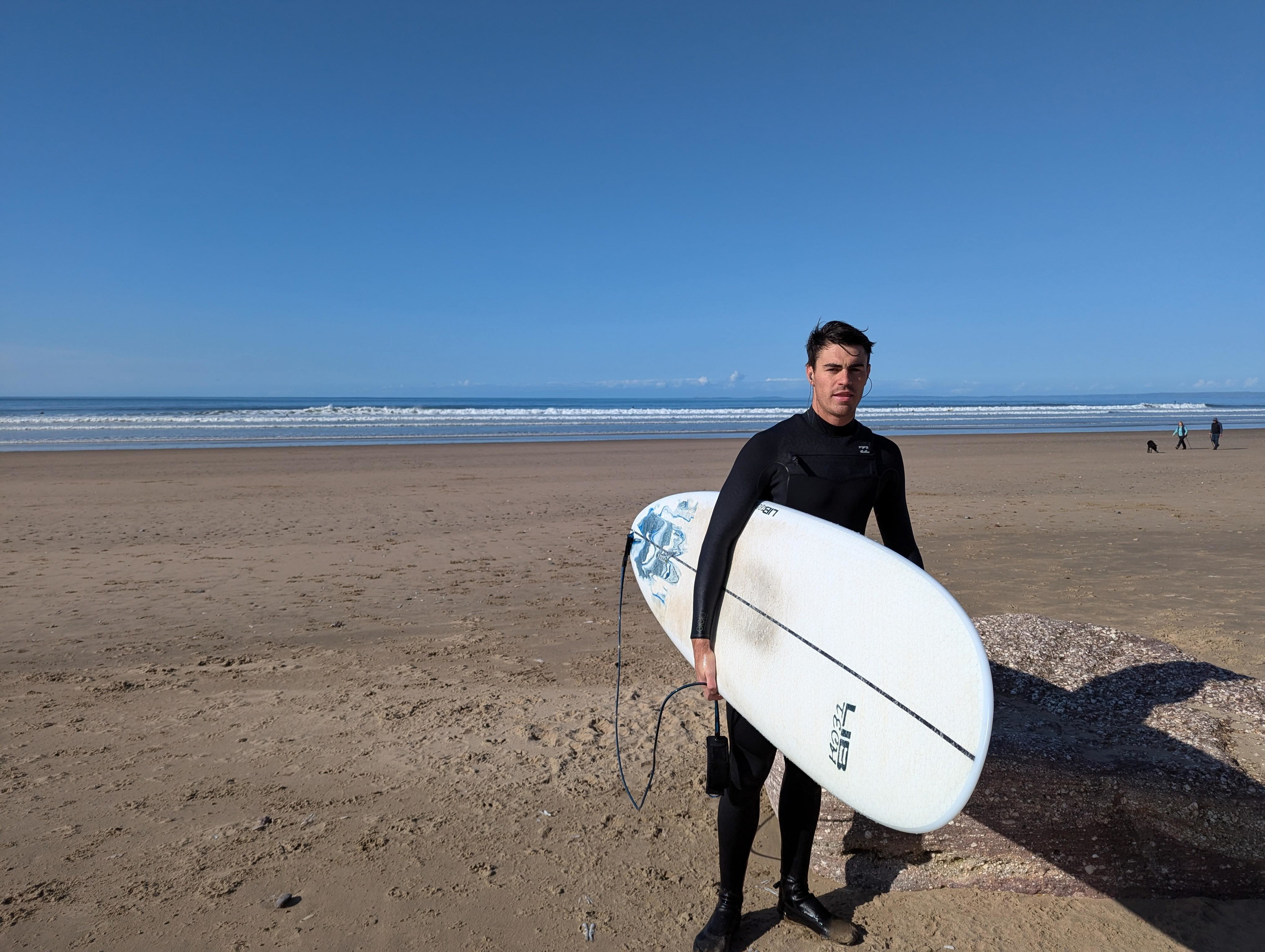 Surfer in a black wetsuit carrying a white shortboard along a wide sandy beach in sunshine