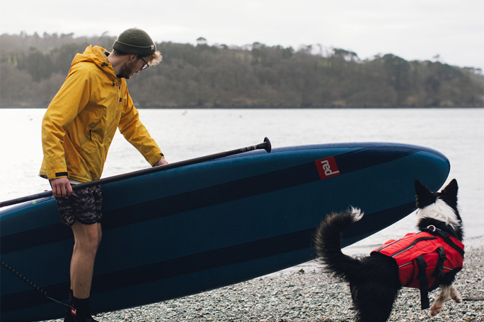 A man carrying a paddle board with his dog