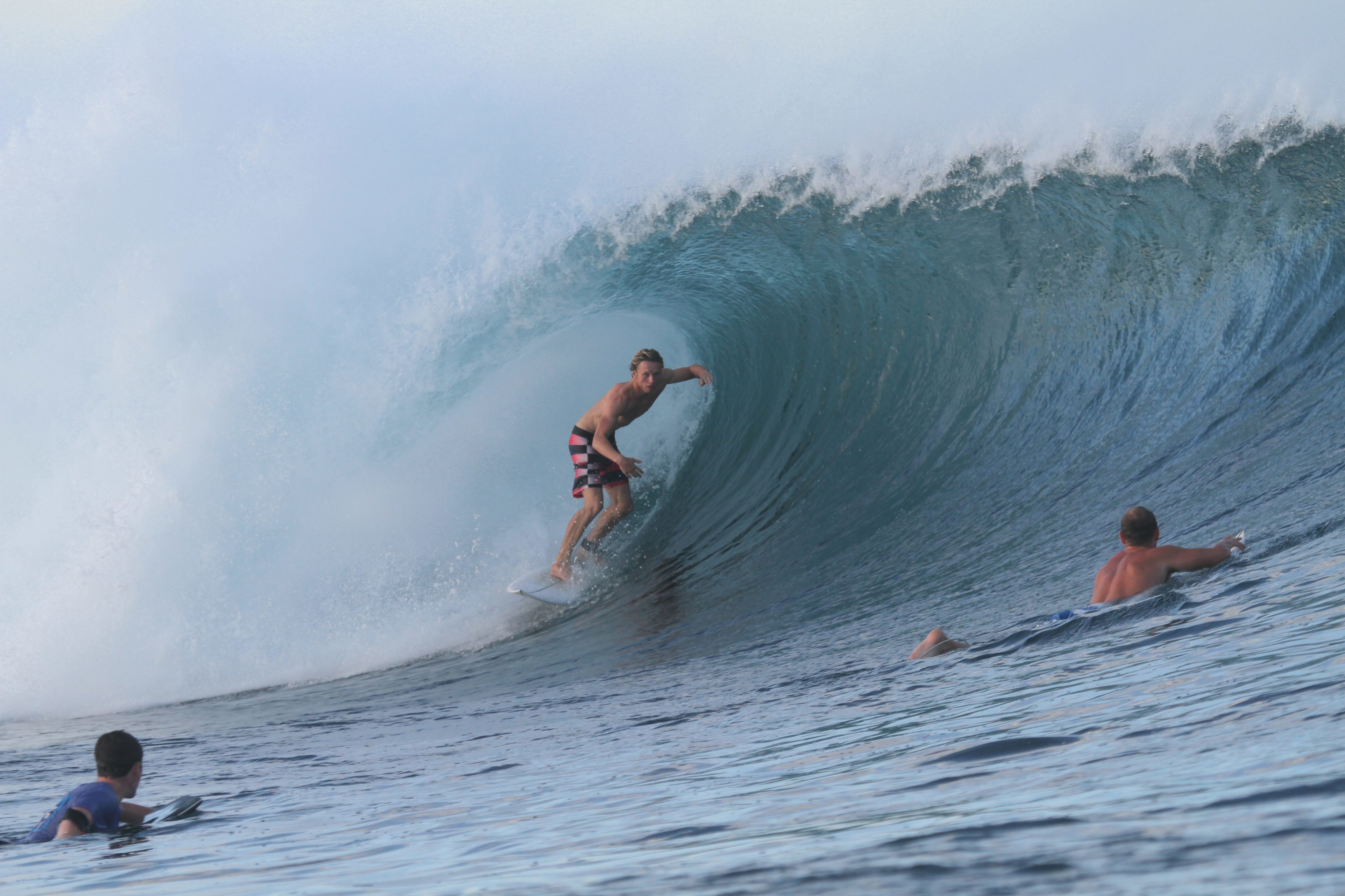 Surfer riding deep inside a large turquoise barrel while others paddle out behind the wave