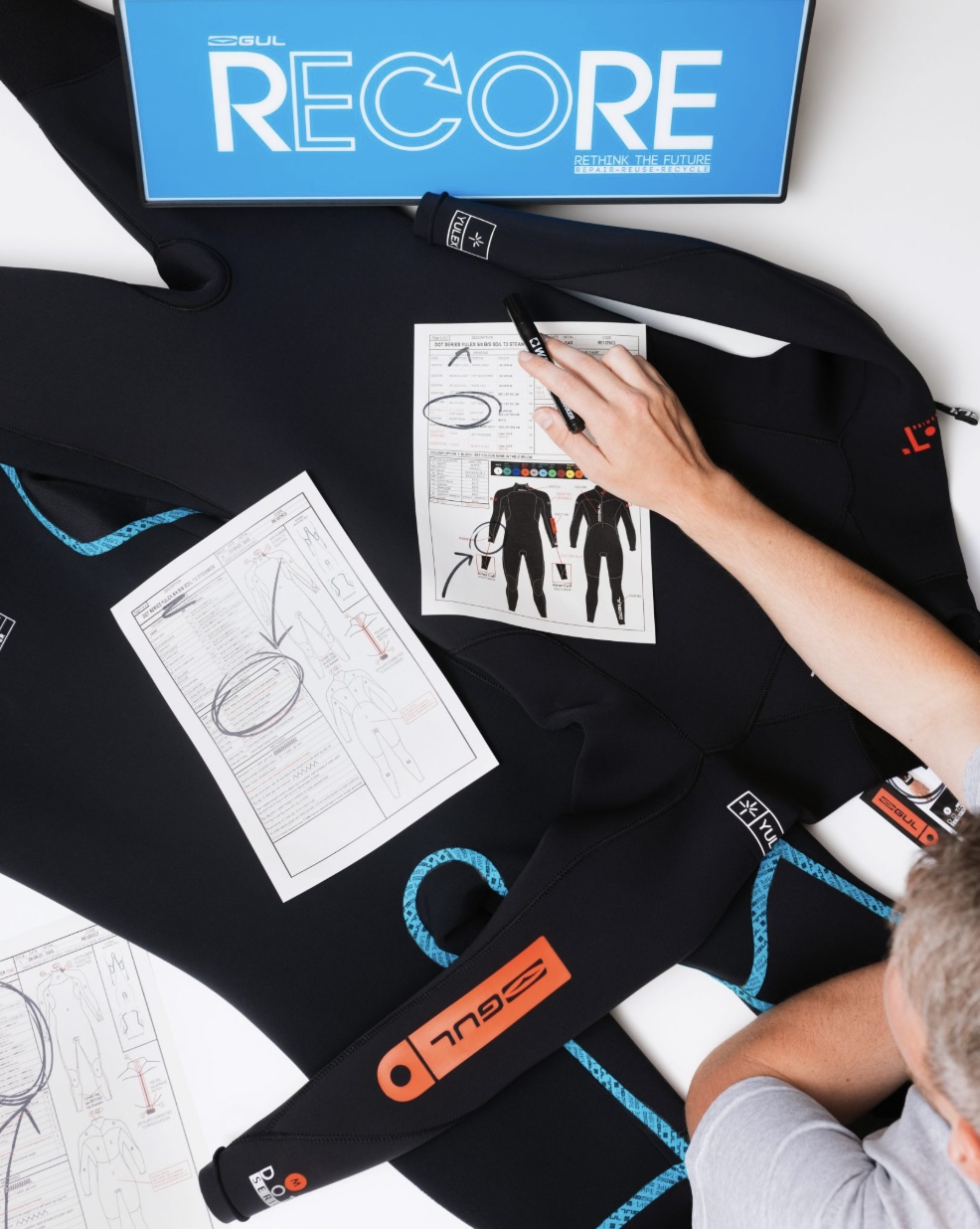 Designer sketching wetsuit drawings on a bench beside a black GUL wetsuit and a RECORE sign