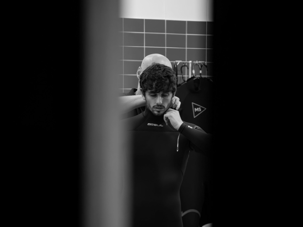Black and white portrait of a young man in a wetsuit and swim cap leaning against a tiled wall