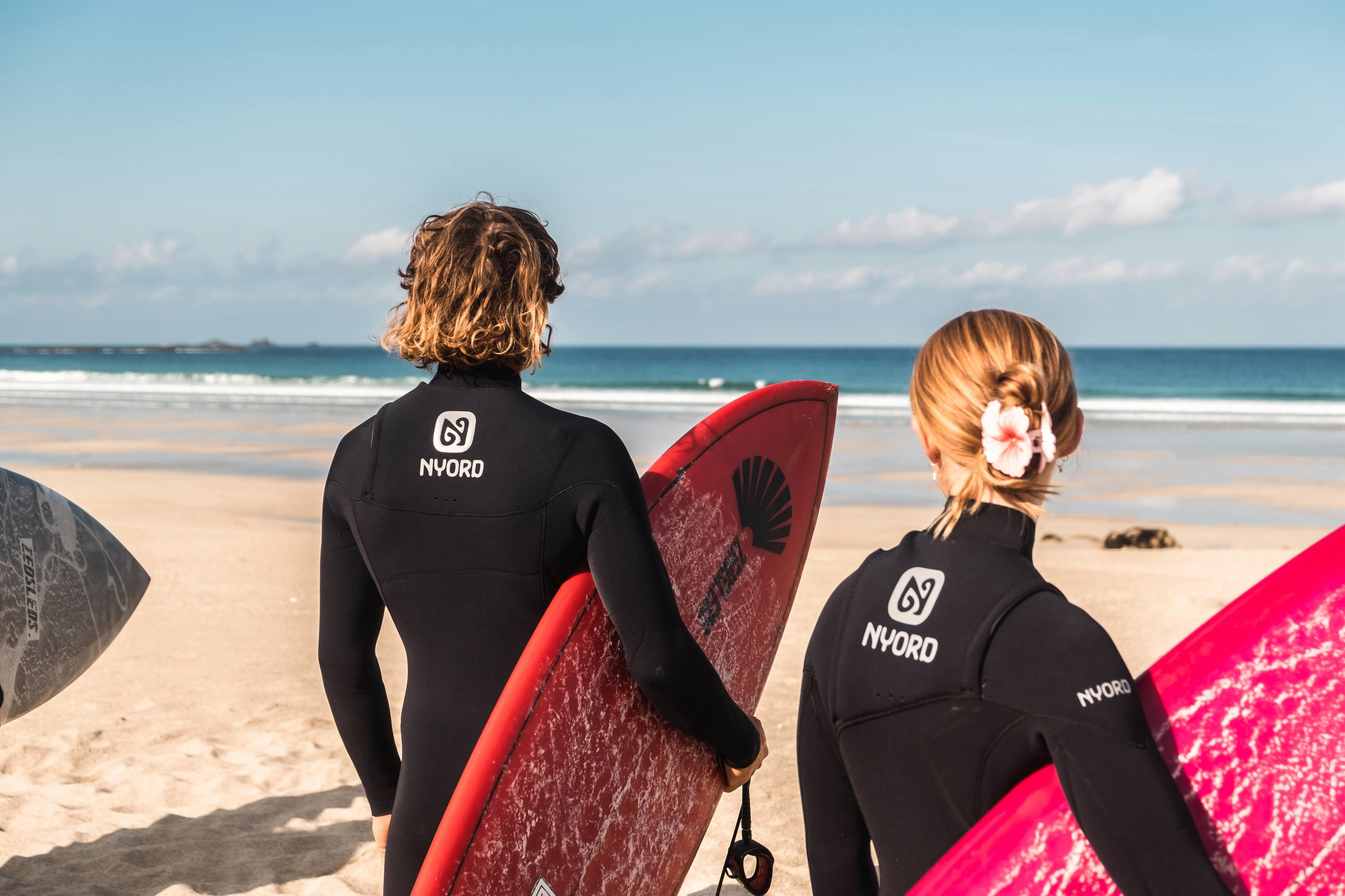 Two surfers in black Nyord wetsuits walking to the sea carrying red and pink boards