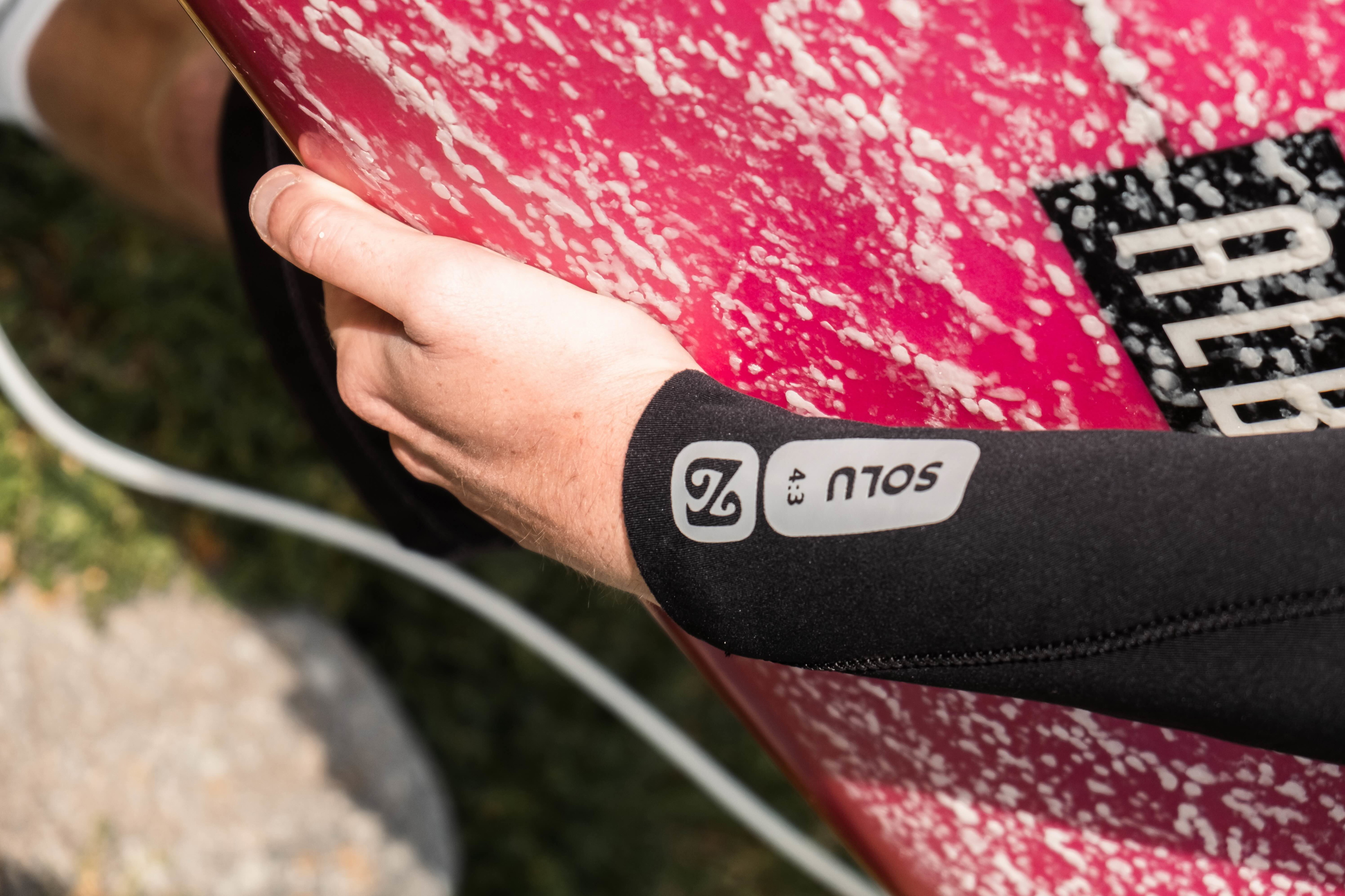 Close-up of a Nyord wetsuit sleeve marked Solu as the surfer holds a pink board underarm