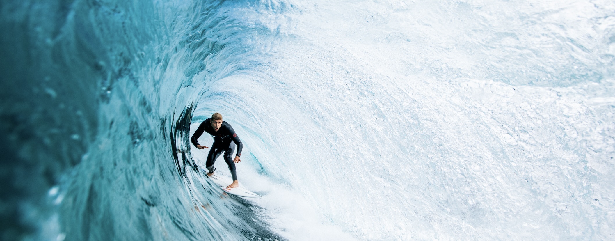 Surfer in a dark wetsuit tucked inside a wide pale blue barrelling wave