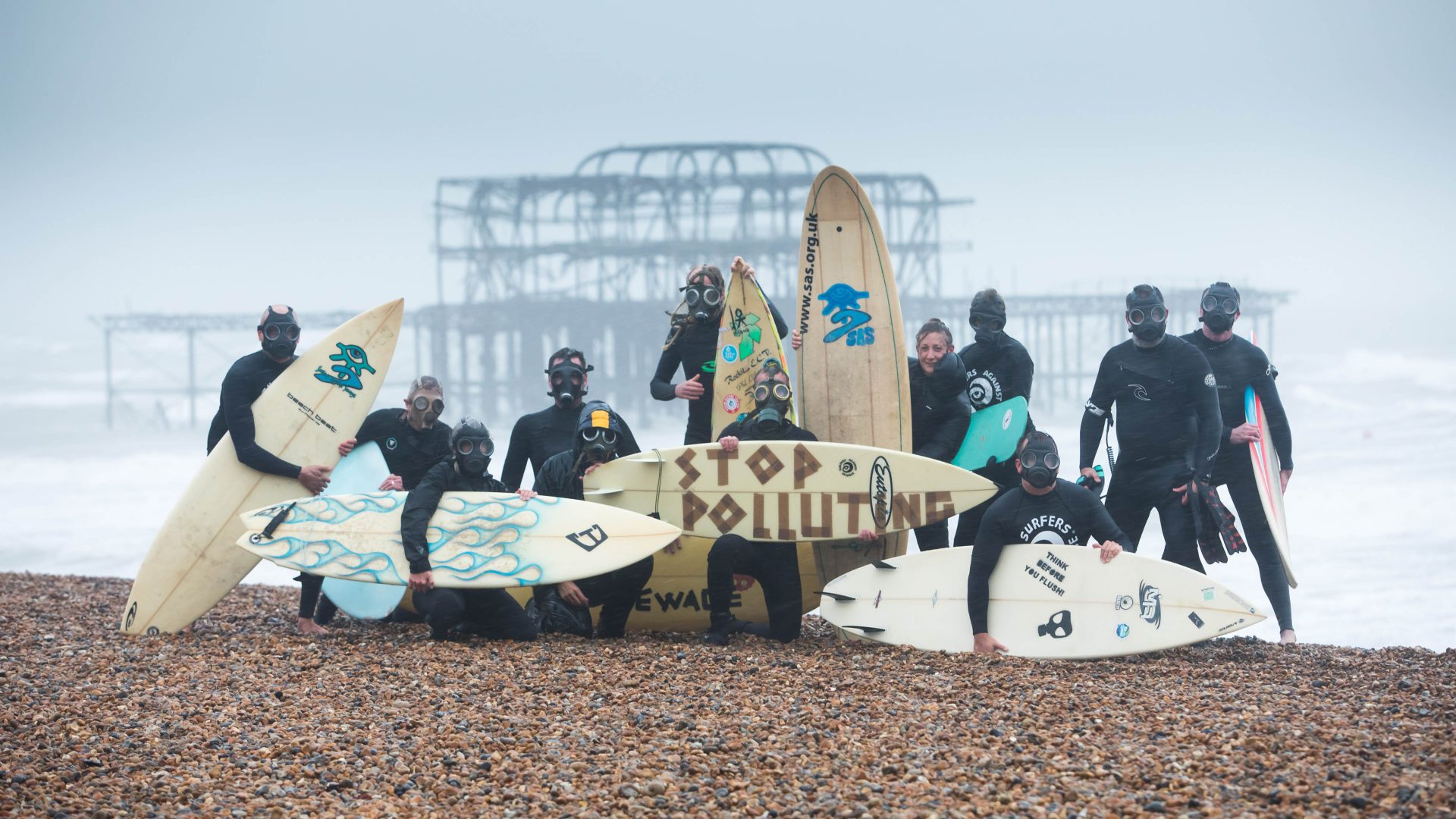 Surfers in wetsuits and gas masks line up with their boards on a shingle beach for a pollution protest