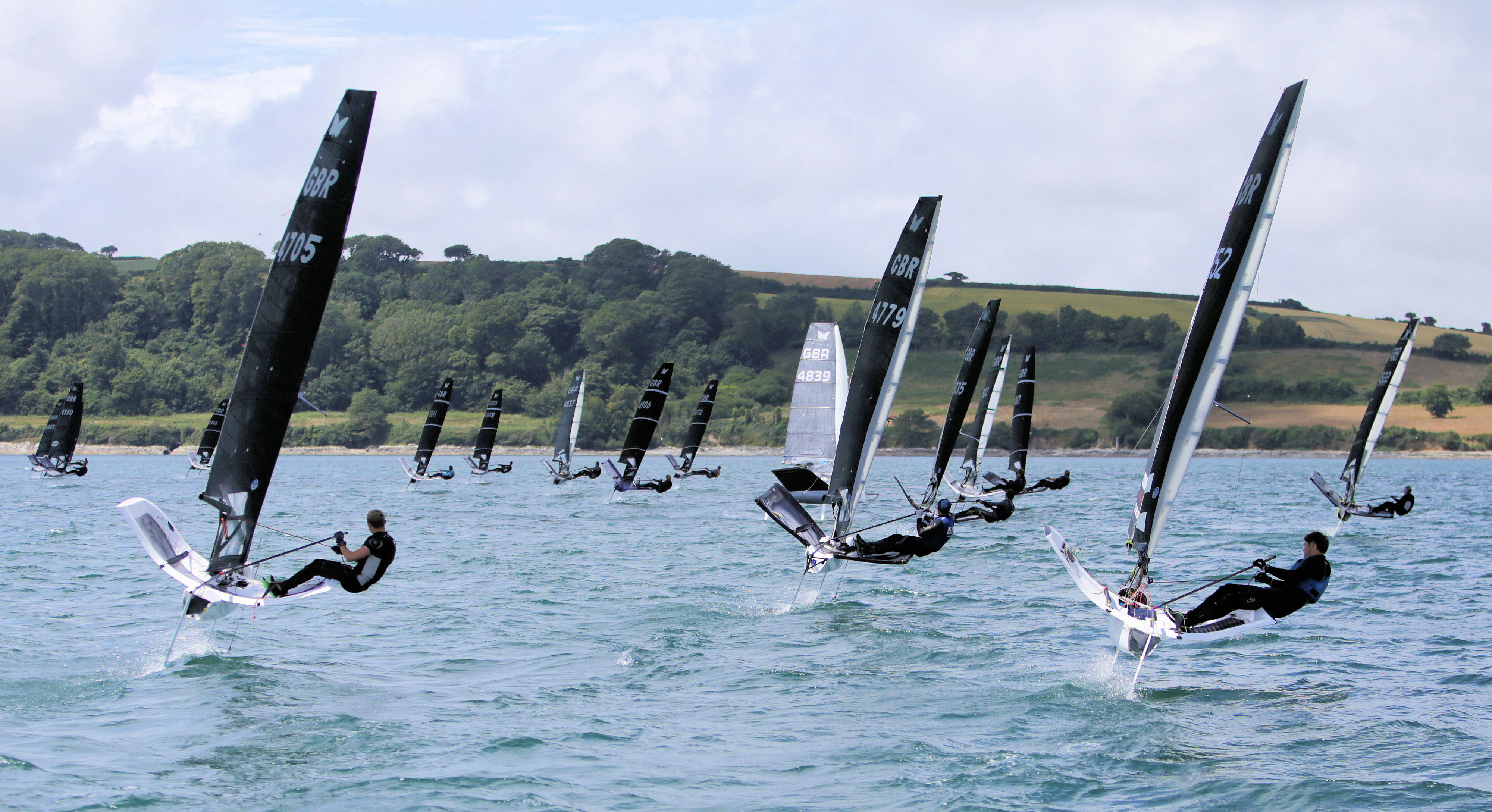 Fleet of Moths foiling away from a race start along a green coastline in bright conditions
