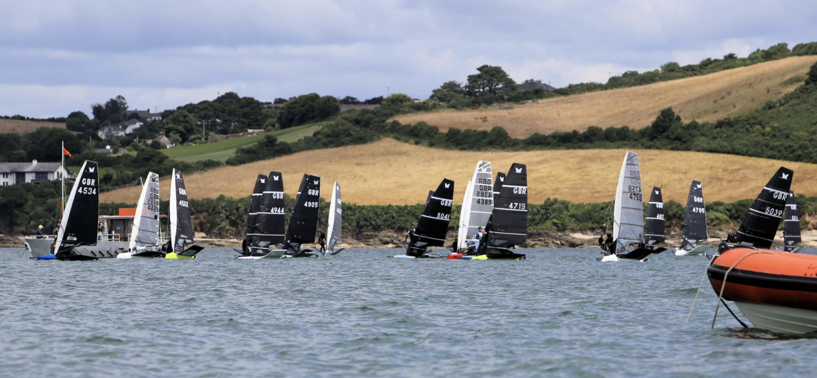 Fleet of Moth dinghies sailing on sheltered water below green hills, with an orange support RIB