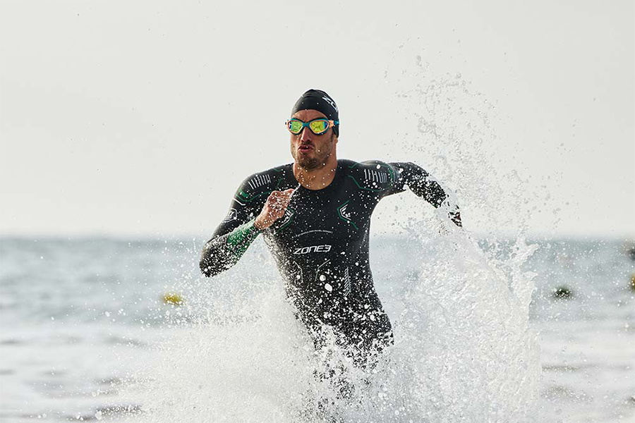 Man in a ZONE3 swim wetsuit wading out through breaking surf, spray all around him