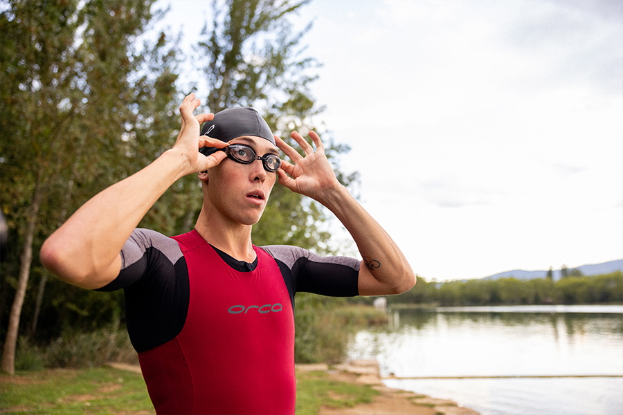 Swimmer in a red and black sleeveless wetsuit adjusting his goggles beside a lake